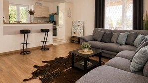 Un salon moderne avec un canapé gris, une table basse et un coin cuisine équipé d’un réfrigérateur, d’un four et de tabourets de bar.