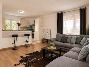 Ein modernes Wohnzimmer mit grauem Sofa, einem Couchtisch und einer Küchenzeile inklusive Kühlschrank, Backofen und Barhocker.
