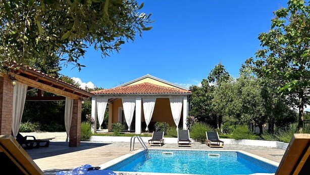 Un espace piscine avec des chaises longues, un coin salon abrité et un bâtiment doté d’un toit en tuiles.