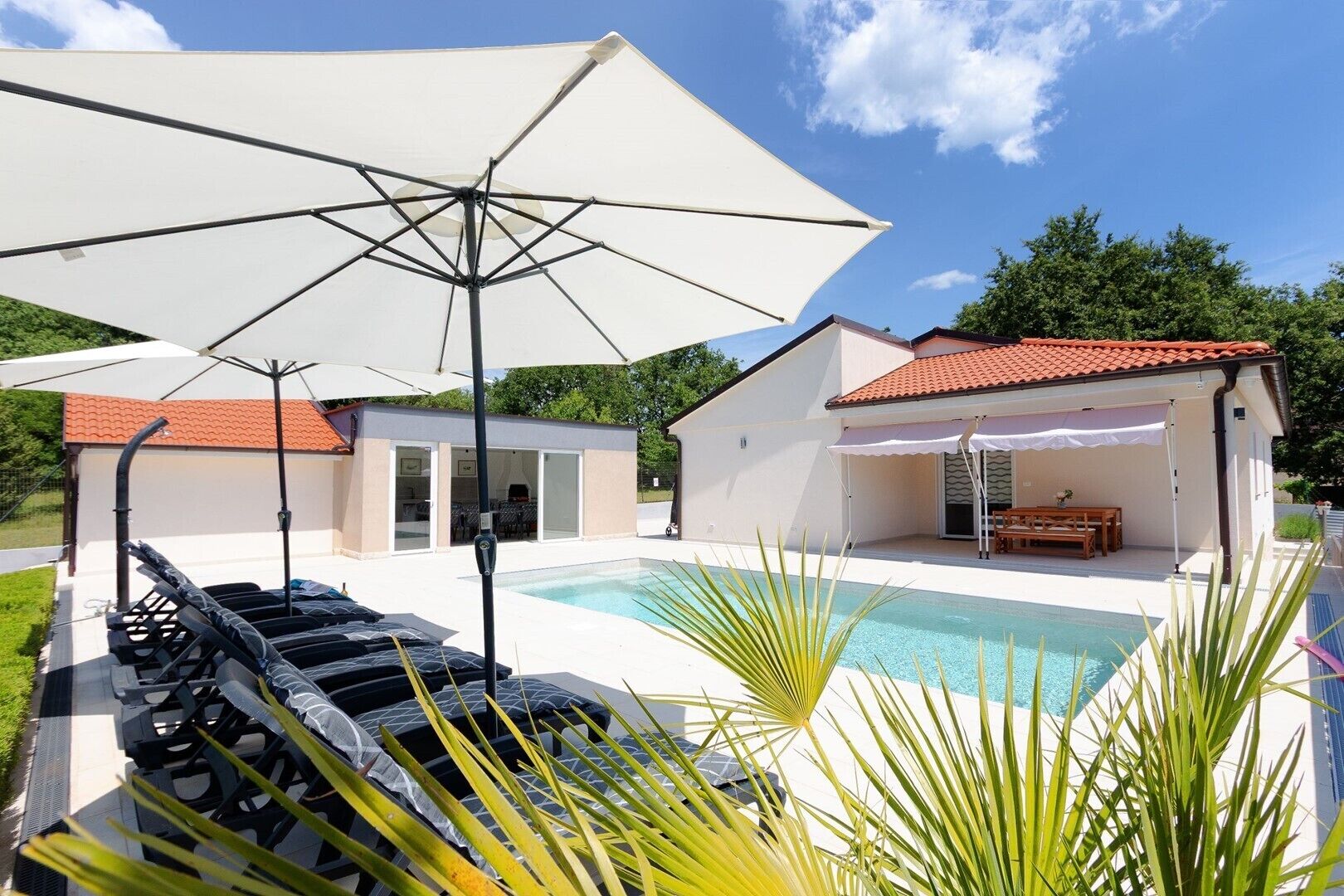 A poolside area with lounge chairs, a large umbrella, and a small building with a red-tiled roof.