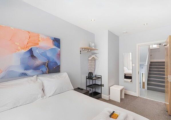 A modern hotel room with a large bed, a painting on the wall, a small table with a remote control, a nightstand, a mirror, and a staircase leading to another room. - Double Room with Private Bathroom - R1 (London)