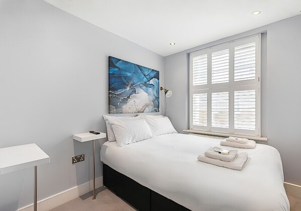 A bedroom with a bed, a nightstand, a painting, a window with blinds, and a wall-mounted light. - Modern Double Room with Ensuite - R3 (London)