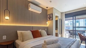 Ett hotellrum med en säng, sängbord, en luftkonditioneringsanläggning, en väggmonterad lampa och utsikt över staden genom stora fönster.