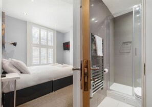 A bedroom with a bed, bedside table, and a window with blinds. - Comfortable Double Room with Ensuite - R4 (London)