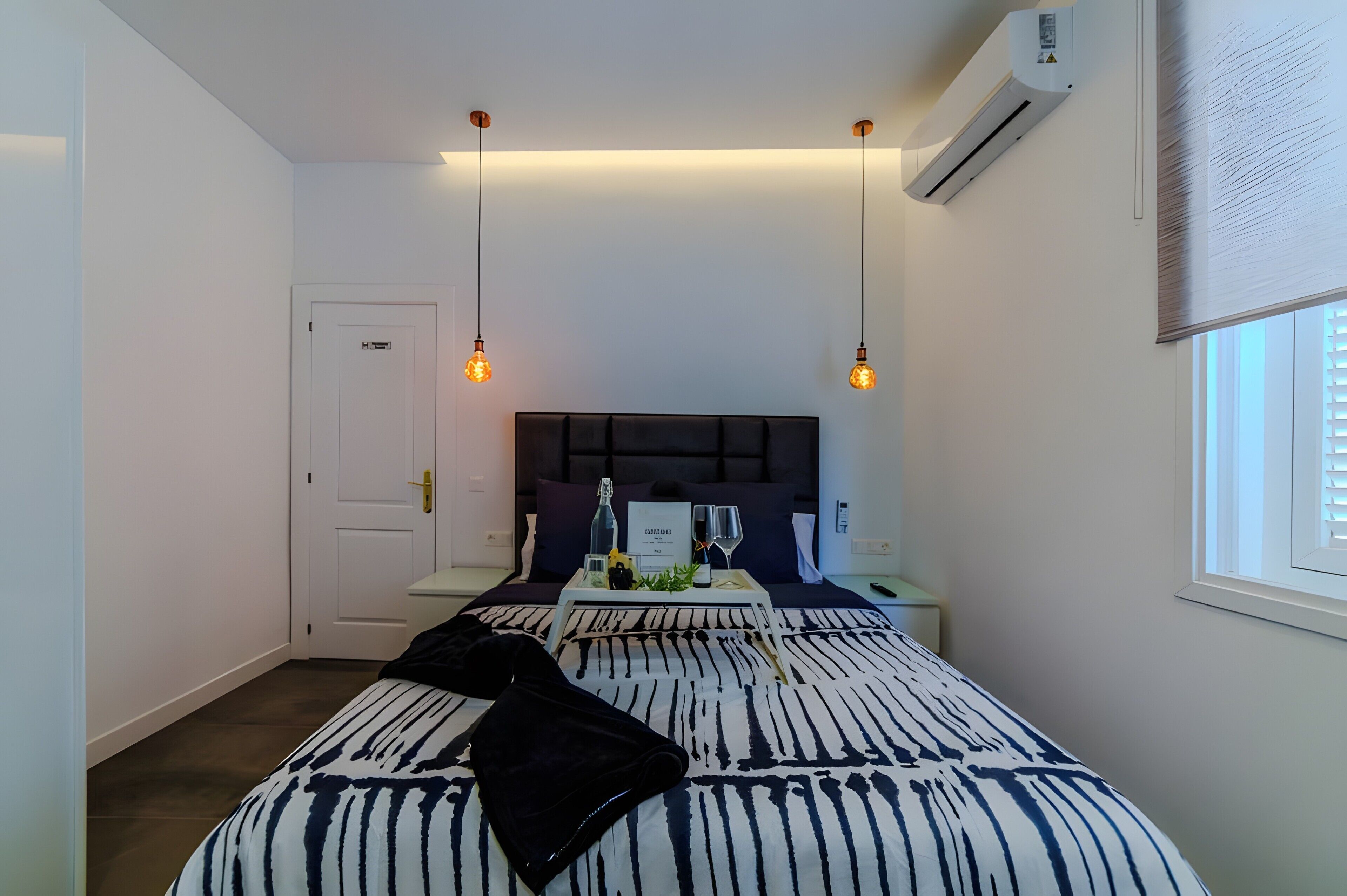 A hotel room with a bed, bedside table, nightstand, and a window with blinds.