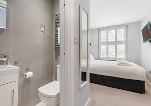 Ein modernes Badezimmer mit Toilette, Waschbecken und Handtuchhalter. Der Raum verfügt über ein großes Bett mit schwarzem Kopfteil und ein Fenster mit Jalousien.