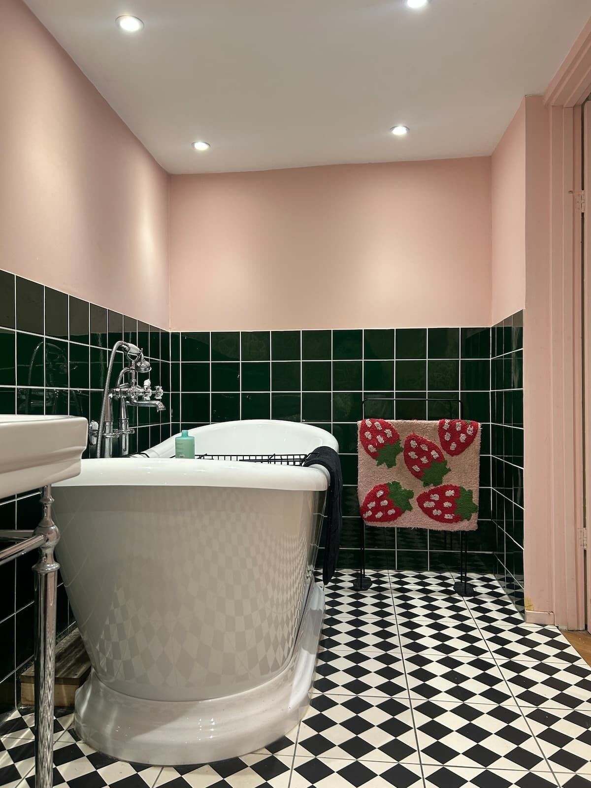 A bathroom with a white clawfoot bathtub, a sink, and a towel rack with a strawberry-patterned towel. The walls are tiled in green, and the floor has a black and white checkered pattern.