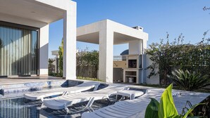 Un área de piscina moderna con sillones de descanso, un cielo azul claro y un edificio con grandes ventanales y un patio cubierto.