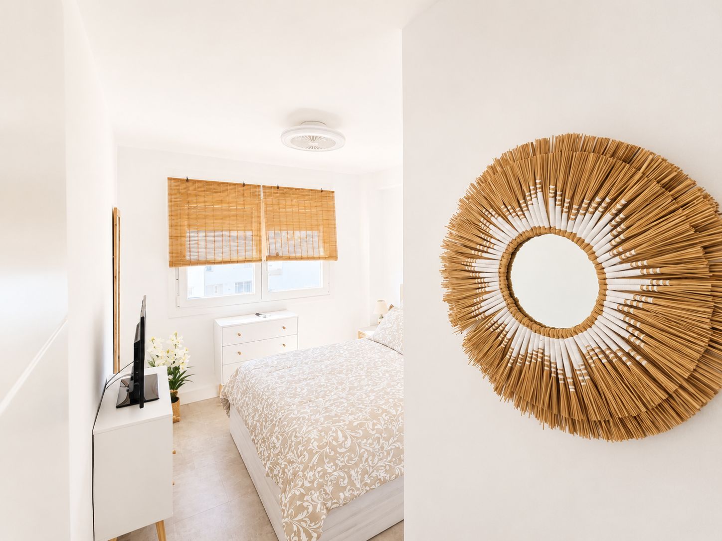 Une chambre à coucher comprenant un lit, une table de chevet, une commode, une fenêtre avec des stores et un grand miroir rond fixé au mur.
