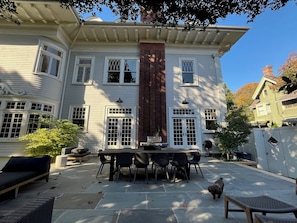Ein großes Haus mit einer Terrasse, auf der ein Esstisch und Stühle stehen, ein kleiner Hund und ein Garten.