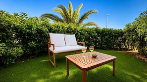 A wooden outdoor seating area with a white cushioned chair and a table, accompanied by two glasses of white wine. - Vera Playa Beachtfront Apartment with garden & 4 Pools (Vera Playa)