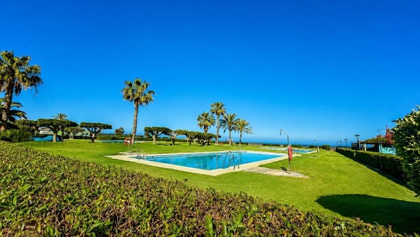 A swimming pool surrounded by well-maintained grass and palm trees, with a clear blue sky. - Casa Vega Vera Playa | Primera Línea de Playa | Beachtfront Apartment with garden & 4 pools (Vera Playa)