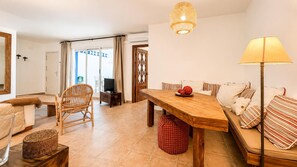 Un salon comprenant une table à manger en bois, un canapé, un fauteuil en osier, un meuble pour la télévision et une lampe.