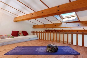 Ein Zimmer mit schrägem Dach, Holzbalken, einem Fenster, einer Couch mit Kissen und einer Yogamatte.