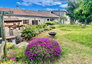 Un bĂątiment rustique en pierre, surmontĂ© dâun toit de tuiles, entourĂ© dâun jardin bien entretenu oĂč sâĂ©panouissent des fleurs dâun violet Ă©clatant, et agrĂ©mentĂ© dâun grand bassin en mĂ©tal.