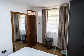 Ein Zimmer mit einem Bett, einer Holztür, einem Fenster mit Vorhängen, einem Mülleimer und einem Kleiderschrank.