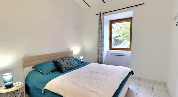 A bedroom with a bed, a nightstand, a lamp, a window with curtains, and a radiator.