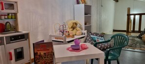 A play kitchen with a toy stove, a toy oven, and a toy sink. There is a green chair and a white table with a purple bowl and a pink cup.