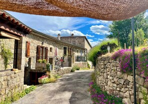 A narrow stone pathway leading to a rustic stone building with a balcony, surrounded by greenery and flowers.