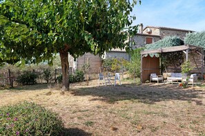 A rustic outdoor seating area with a pergola, a stone building, and a fence in the background.