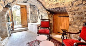 A stone-walled room with a wooden door, a red upholstered chair, a small round table, and a patterned rug. - Guest Room “L’Adrienne” with Shared Garden and Wi-Fi (Chazeaux)