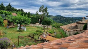 A rustic stone building with a terracotta roof, surrounded by lush greenery and a clear sky.