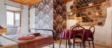 A room with a bed, a table with chairs, a stone wall, and a wooden ceiling.