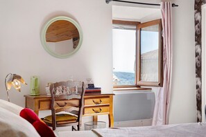 A room with a wooden dresser, a chair, a bed, a round mirror, and a window with curtains.