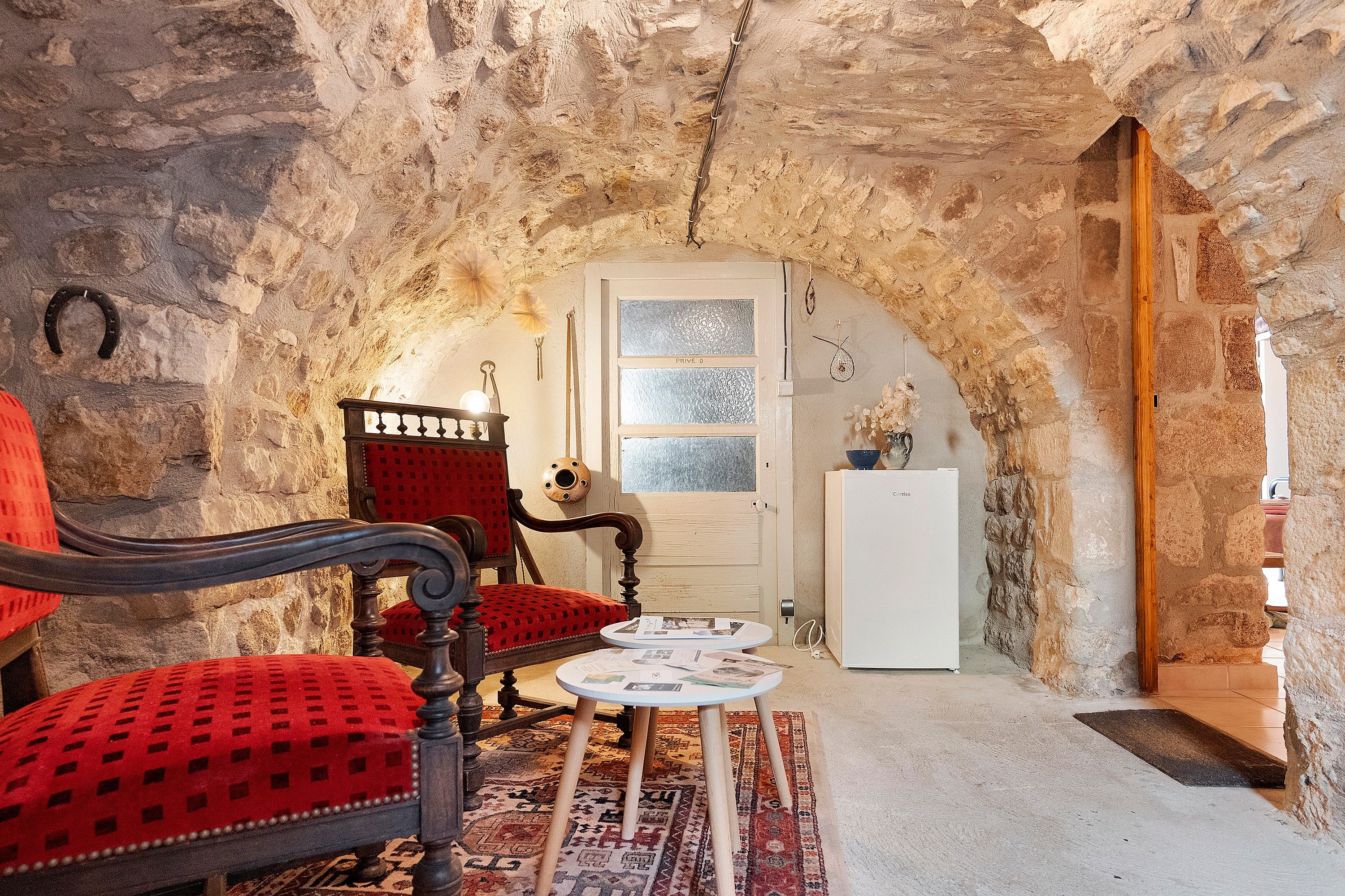 Quarto rústico com paredes de pedra, porta de madeira, poltrona vermelha estofada, mesa pequena com papéis e geladeira branca.