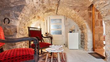 Quarto rústico com paredes de pedra, porta de madeira, poltrona vermelha estofada, mesa pequena com papéis e geladeira branca.
