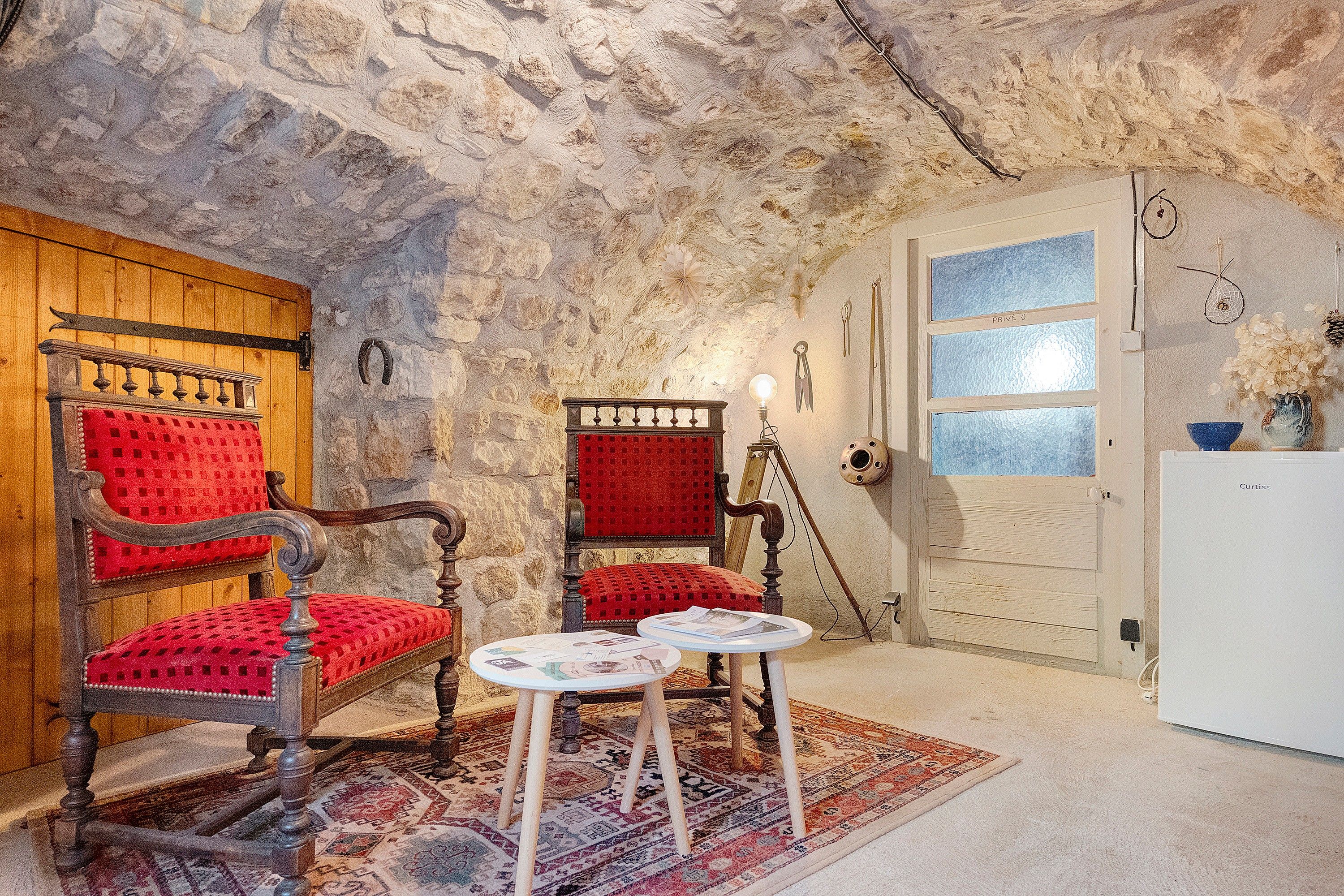 Une pièce avec un mur en pierre, une porte en bois, un fauteuil rouge recouvert de tissu, une petite table avec des papiers et un réfrigérateur blanc.