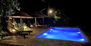 Una piscina nocturna con sillones de descanso y sombrillas.