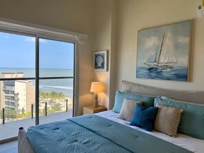 Una camera da letto con una grande finestra, un letto, un comodino, un quadro e vista sull'oceano.