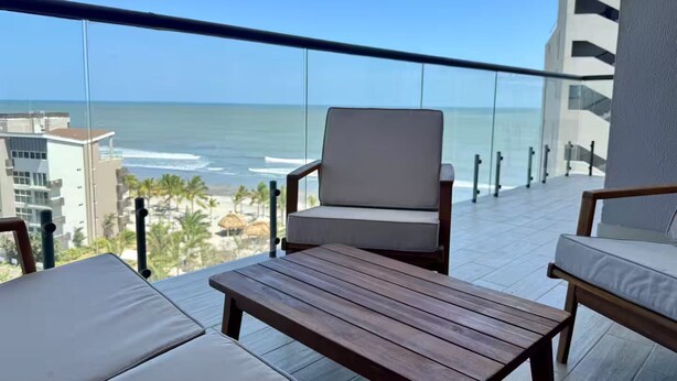 Un balcón con mesa y dos sillas de madera, con vista a una playa y al océano.