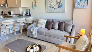 Una sala de estar con un sofá gris, sillas de madera, una mesa de centro con objetos decorativos, una zona de cocina y obras de arte en las paredes.