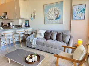 A living room with a grey sofa, wooden chairs, a coffee table with decorative items, a kitchen area, and artwork on the walls. - Apartamento con increíble vista frente al mar (punta chame)
