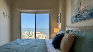 Un dormitorio con un ventanal grande, una cama, un cuadro y vistas a edificios y al agua.