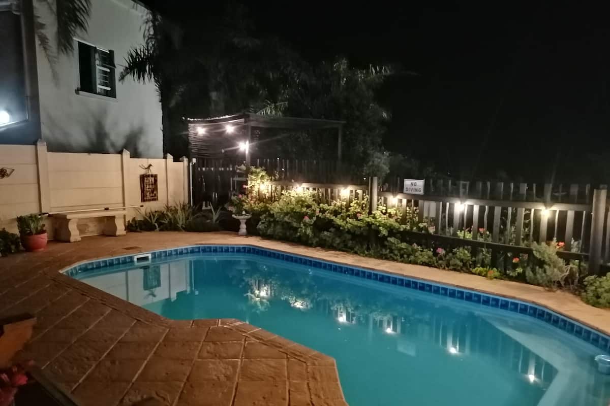 A nighttime swimming pool with a tiled edge, surrounded by a wooden deck and potted plants.