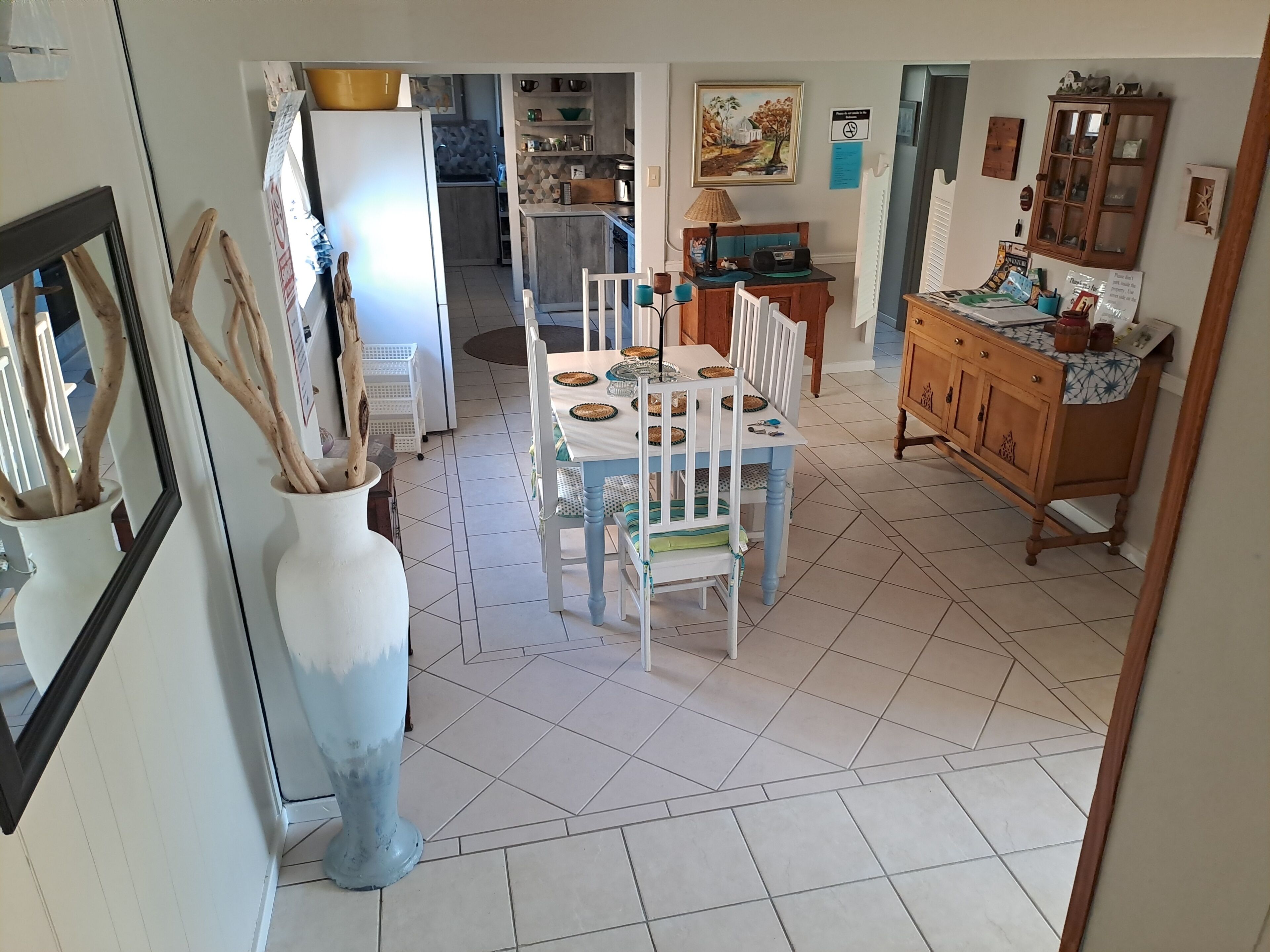 Un comedor con una mesa dispuesta para dos personas, una cocina al fondo y un jarrón grande con ramas.