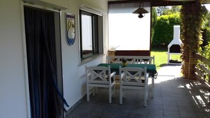 Une terrasse avec une table et des chaises, une fenêtre avec des rideaux, une enseigne fixée au mur et un barbecue à l’extérieur.