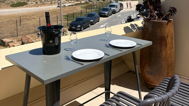 A balcony with a table set for two, a potted plant, and a view of the sea and vehicles in the background. - Naturist Village - Heliopolis AB - 30 m² - ocean view (Agde)