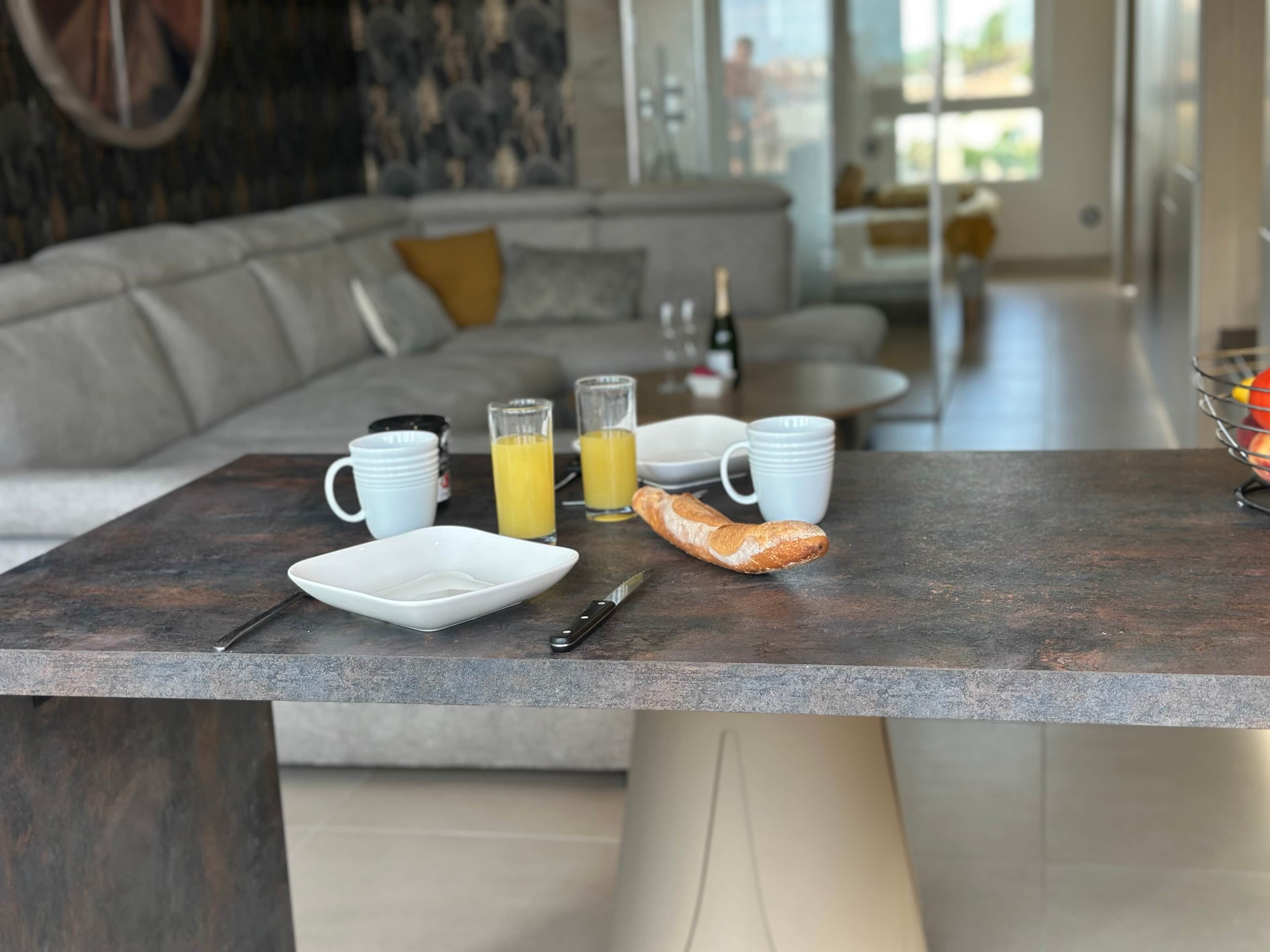 A modern kitchen with a dining table set for breakfast, including a baguette, glasses of orange juice, and white cups.