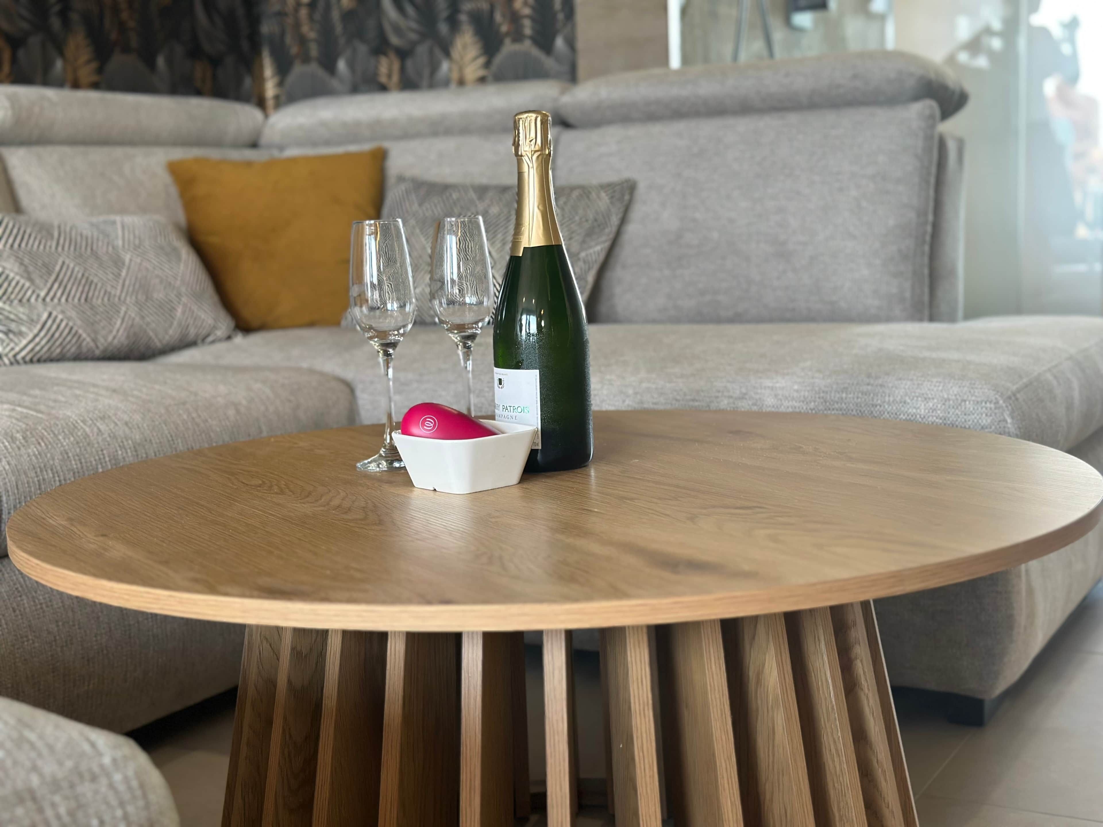 A round wooden table with a bottle of champagne, two glasses, and a white bowl with a pink object.