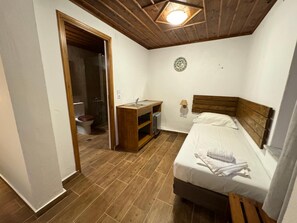 Ein Schlafzimmer mit hölzernem Kopfteil, einem Einzelbett, einem Nachttisch, einem kleinen Schreibtisch mit Stuhl, einer Toilette und einem Duschbereich.