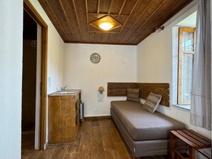 Ein gemütlicher Raum mit Holzbalkendecke, einem Bett mit Kopfteil, einem kleinen Holzschrank, einer Wandleuchte und einem Fenster mit Vorhängen.