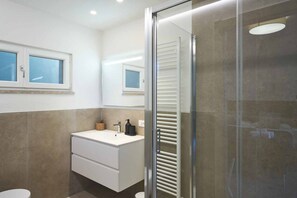 Un baño moderno con cabina de ducha de vidrio, un tocador blanco con lavamanos y una ventana sobre el espejo.