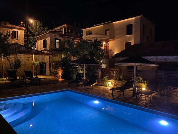 A swimming pool with lounge chairs and umbrellas, surrounded by stone buildings at night. - Tsagarakis Guesthouse & Pool Room.2 with Mountain & Pool View, Wi-Fi, and Air Conditioning (Xinovrysi)