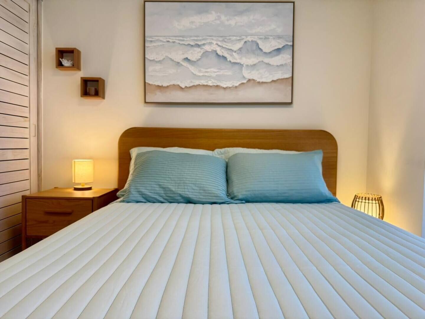 A bed with a striped white and blue bedding, a wooden headboard, a bedside table with a lamp, and a framed picture of waves on the wall.