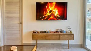 Quarto com uma cama, televisão na parede exibindo uma lareira, mesa consola de madeira com objetos decorativos e uma porta.