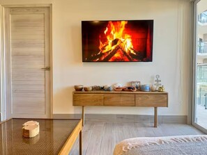 A room with a bed, a TV mounted on the wall displaying a fire, a wooden console table with decorative items, and a door. - Apartamento frente al mar con vista a la piscina (punta chame)
